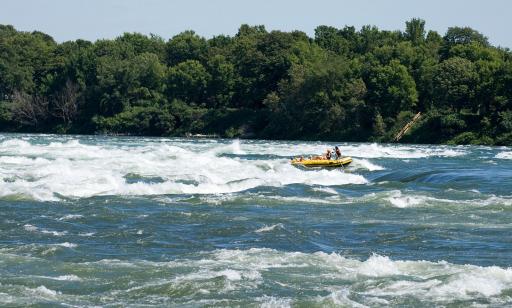 People ride an inflatable boat on turbulent rapids, not far from a tree-covered island.