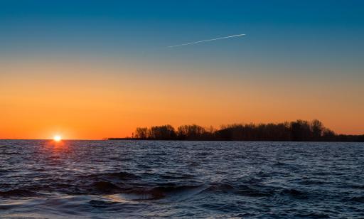 Trees on an island are silhouetted against the bright orange sky as the sun breaks the horizon over the water.