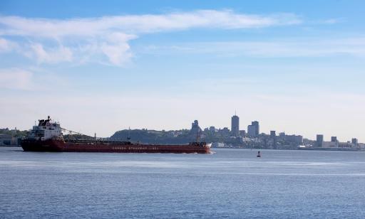 Un cargo rouge navigue vers l’aval sur le Saint-Laurent et on voit au loin les silhouettes du Cap Diamant et des gratte-ciel.
