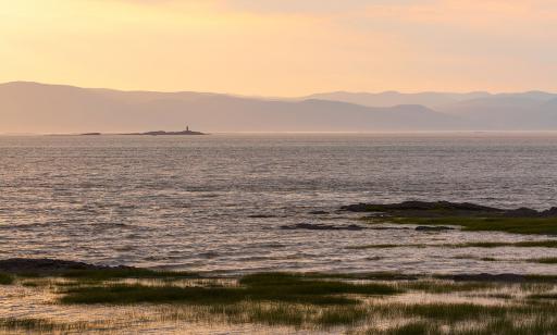 Une légère brume enveloppe les silhouettes lointaines des montagnes et d’un phare sur un îlot rocheux au milieu du fleuve.