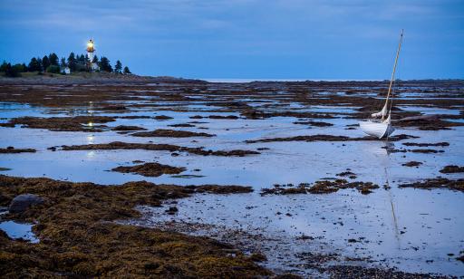 A sailboat sits on the shoreline at low tide. In the twilight, a distant lighthouse shines out from a rocky point.