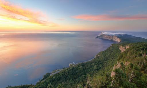 Lumière rosée du soleil levant au-dessus de la vaste étendue d’eau du golfe, des falaises de roc et des forêts touffues.