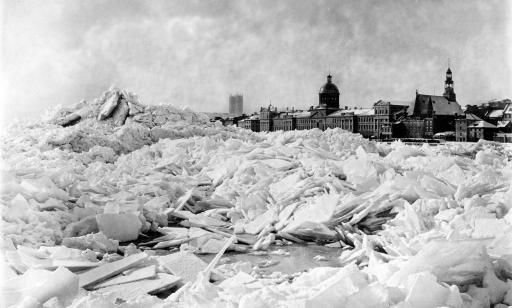 Large mounds of ice piled up on the water with church steeples and other buildings in the background. 