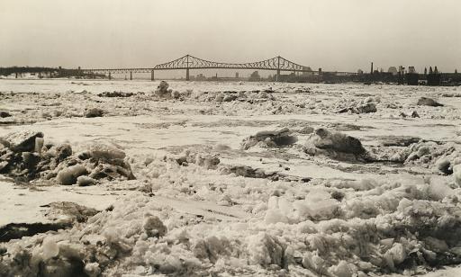 Amoncellement imposant de glaces sur l'eau et silhouette d’un pont et des arbres sur les rives se détachant sur l’horizon.