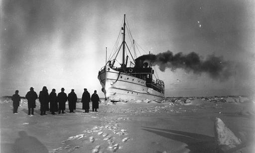 Eight men stand on the frozen river in front of an icebreaker. Thick black smoke billows from the ship's stack.