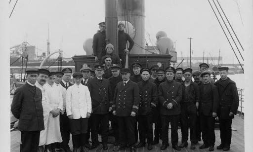 Vingt-trois hommes vêtus d’un costume de marin et quatre hommes vêtus de blanc se tiennent debout sur le pont d’un navire.