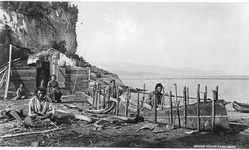 Six Aboriginal people on a shoreline. Two of them use stakes to shape a sheet of birchbark into a canoe.