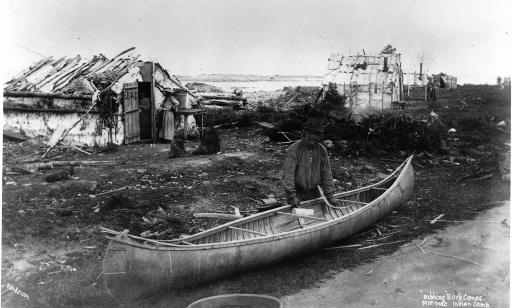 An Aboriginal man on a shoreline, in front of three cabins, adds a curved strip of wood to the ribbing of a canoe.