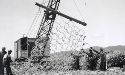 Men tie cables to a wide-mesh metal net, part of which has been lifted by a crane.