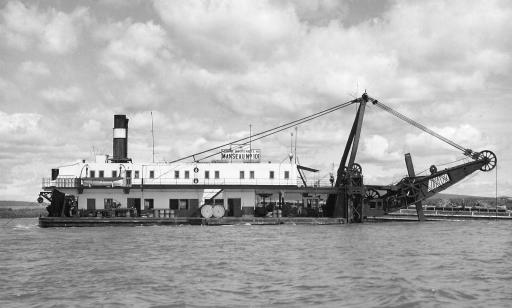 Black and white photo showing a side view of a dredge, with dredging equipment at the front.