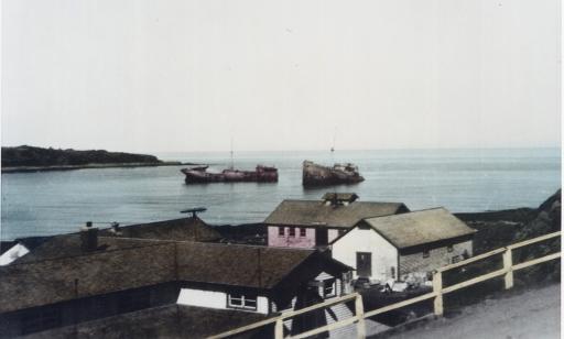 Lightly colorized black and white photo of a wreck split into two pieces lying on the bottom of a shallow bay.