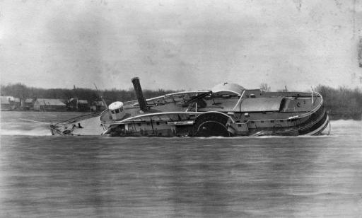 Un bateau à vapeur à roues à aubes échoué et incliné à bâbord dans l’eau. On voit des habitations au loin sur la berge.