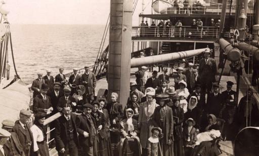 Several men, women and children stand on the decks of a ship, while two women remain seated.