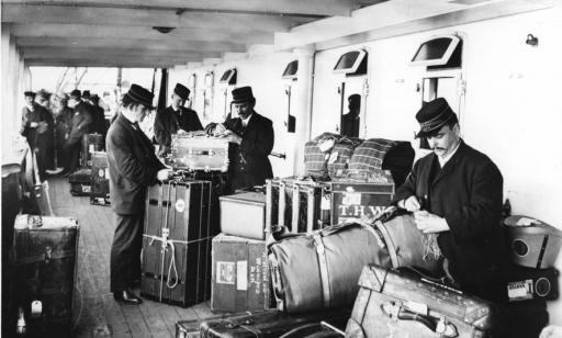 Men wearing caps and uniforms place tags on various pieces of luggage piled up on the deck.