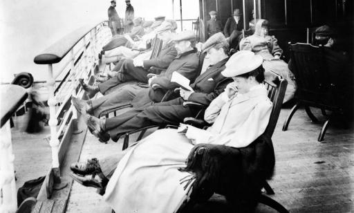 Des femmes et des hommes lisent assis sur des chaises longues en bois sur un pont d’un paquebot du début du XXe siècle. 