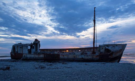 Light from the setting sun shines through cracks in the wreck of a wooden trawler lying on the shore.