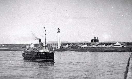 A boat with a single smoke stack sails near the shore, in front of a lighthouse, a church, houses and other buildings.