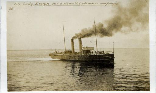 Two sailors stand on the deck of a boat in calm waters. Its two smoke stacks emit clouds of dark smoke.