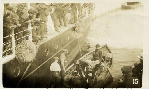 A man in a rowboat prepares to move the body of a victim to a ship, where men stand ready to receive it. 