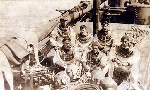 Six men in diving suits seated on the deck of a ship, with their helmets in front of them.