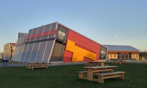 Grey corrugated metal building with red and yellow accents. Part of the building suggests a ship leaning on its side.