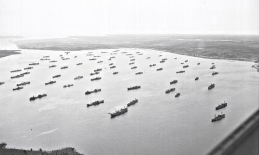 Aerial view of many ships grouped together on the water. Almost all are facing in the same direction.