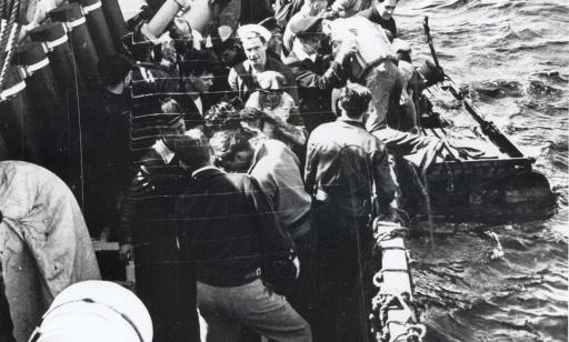 Several men on the deck of a corvette help four men out of a fragile raft.