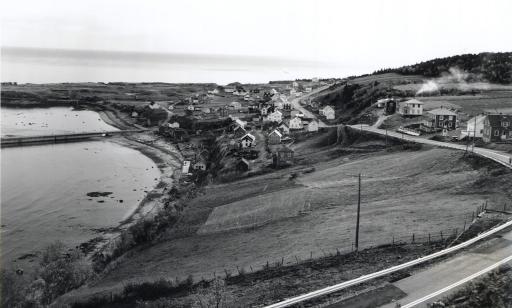 Photo en noir et blanc prise à partir du haut d’un escarpement montrant un village sur le bord du golfe du Saint-Laurent.