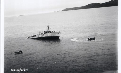 Deux chaloupes sont près d’un navire de guerre incliné à bâbord dans une baie après une collision avec un autre navire.