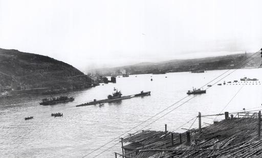 Un sous-marin allemand entouré de navires et de chaloupes se trouve dans une baie bordée d’un paysage montagneux.