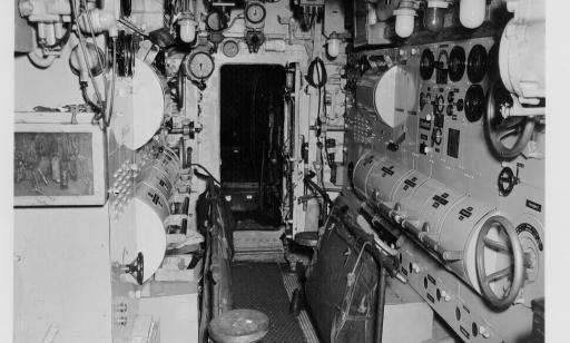 Black and white photo showing controls, valves, lights and dials, as well as a door and two stools.