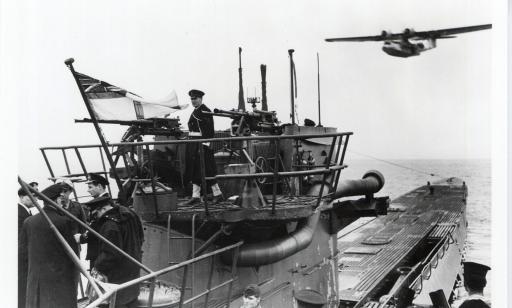 A sailor of the Royal Canadian Navy, wearing a white armband and gaiters, raises the White Ensign on the mast of a U-boat.