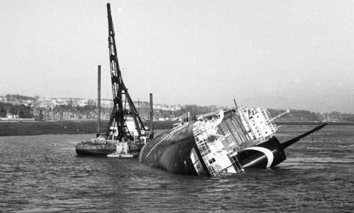 A ship lies nearly keeled over on its starboard side. A crane stands on a barge by its port side. 