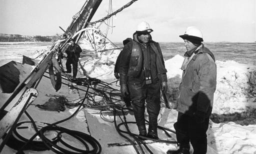 Four men stand on a beached ship, keeled over on its side and covered in snow. A number of cables can be seen.