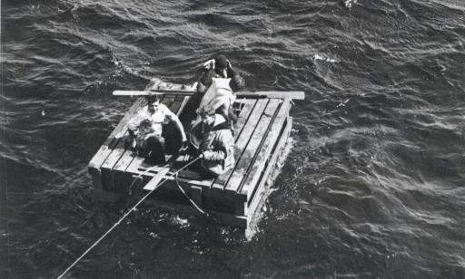 Four men wearing lifejackets on a makeshift wooden raft being towed with a cable.