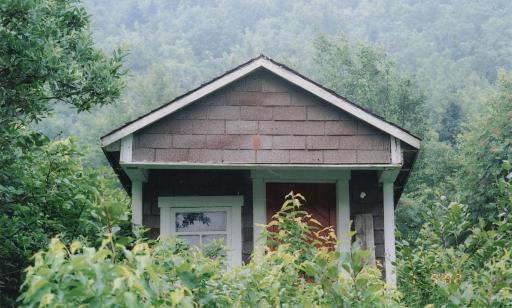 Petite cabane recouverte de bardeaux d’asphalte bruns et gris avec bordures blanches en bois dans un environnement forestier.