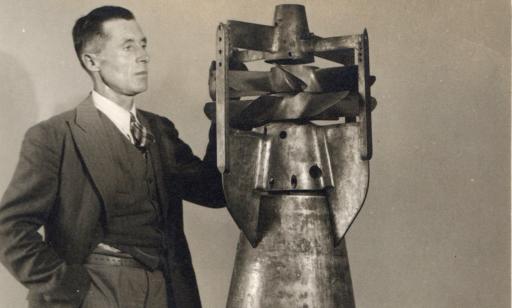 A man wearing a jacket stands with his left hand on a torpedo as tall as he is.