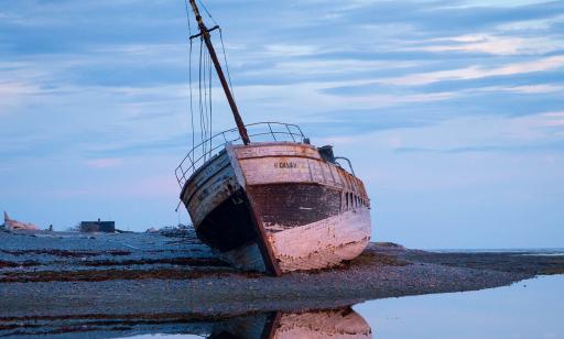 Chalutier en bois échoué sur la berge avec sa réflexion dans l’eau sous un ciel nuageux à la lueur bleutée du crépuscule.