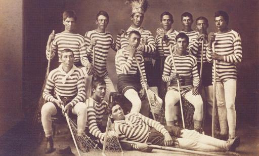 Twelve Mohawk lacrosse players wearing striped sweaters. One man, in the centre back, is wearing a feather headdress.