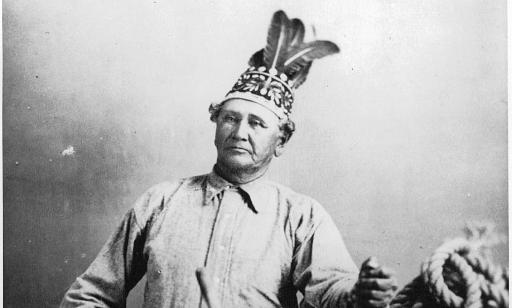 A Mohawk pilot wearing a feather headdress and an arrow sash holds a boat’s wheel in both hands.