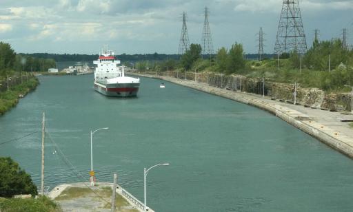 A cargo ship sails along a canal toward the locks. Several electric pylons stand on the shore on its port side.