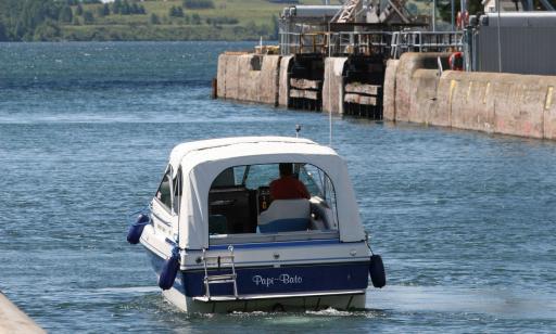 On voit un homme à bord d’un yacht bleu et blanc sortir d’une écluse dont les portes sont ouvertes et le bras d’arrêt levé.