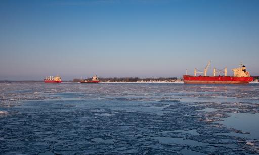 À partir de la rive en hiver, on voit des cargos sur le fleuve partiellement recouvert de glaces devant une île enneigée.