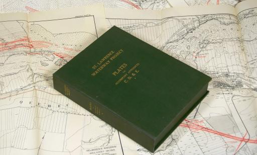 A book with gold lettering on its green cover atop a series of maps showing the route of the St. Lawrence Seaway.