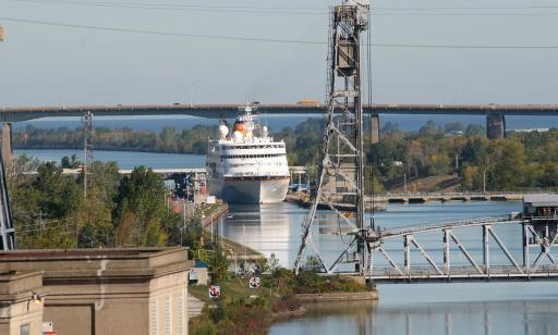 A large white ship heads along a canal toward a lift bridge. The central span is lowered.