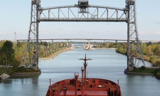 A red cargo ship sails along a canal toward a lift bridge. Its central span is raised.