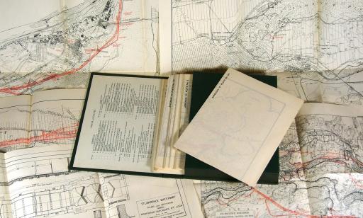 An open book atop maps and plans for a lock. It contains several documents in a pocket.