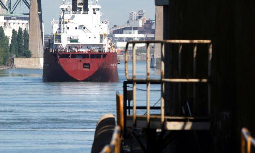 A red and white cargo ship sails out of a lock toward a raised bridge. Port facilities can be seen in the distance.