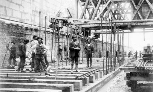 Black and white photo of workers completing various tasks in a trench with concrete-block walls.