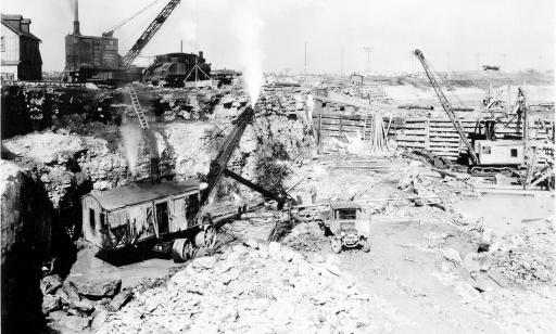 Black and white photo of an excavator in a deep trench. A crane stands at the top by the edge.